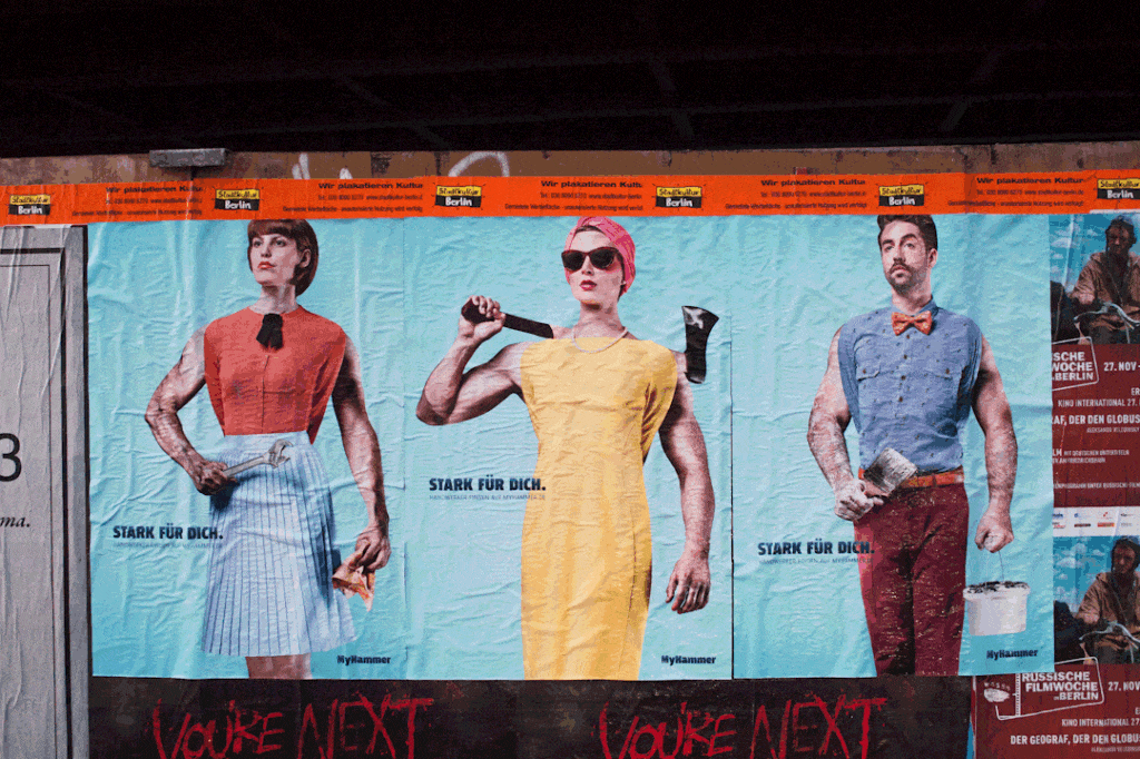 Site fence posters on which the arms of tradesmen holding tools are stretching out from behind different people